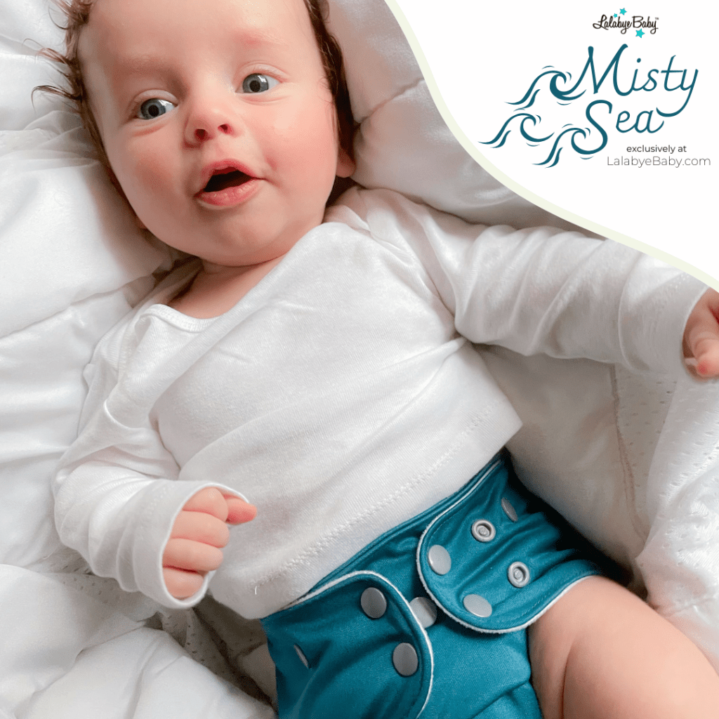 A beautiful caucasian baby around 6 months old with grey eyes, curly brown hair, cherub cheeks, and pink lips lays against a soft white blanketed background. The baby wears a crisp white long sleeved shirt and the new Misty Sea cloth diaper. The color is a deep, gemstone teal: vibrant and luxuriously looking. The diaper has silver snaps.